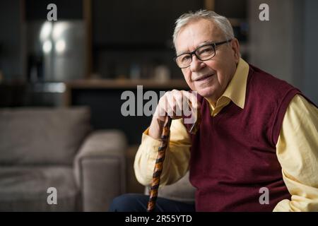 Ein glücklicher Seniorenmann sitzt in seinem Haus auf dem Bett. Porträt eines Seniorenmannes mit Gehstock. Stockfoto