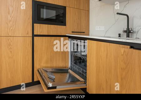 Moderne Küche mit leerer Einbauspülmaschine mit geöffneter Tür, Edelstahl-Spülbecken und Leitungswasser, schwarzem Induktionskochfeld, Herd Stockfoto
