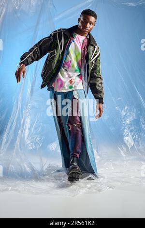 Trendiger junger afroamerikanischer Mann in einer Outwear Jacke mit LED-Streifen und gerissenen Jeans, der läuft und auf glänzendes Zellophan auf blauem Hintergrund auf die Kamera schaut Stockfoto