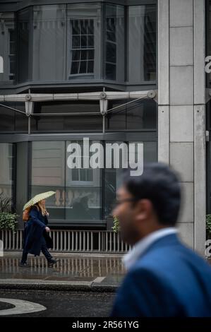 London, Vereinigtes Königreich: Eine Frau läuft im Regen eine Straße entlang und trägt einen Regenschirm. Ein verschwommener Mann ist im Vordergrund und geht in die andere Richtung. Die Stadt London. Stockfoto