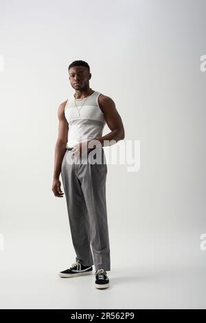 Ein junger afroamerikanischer Mann in ärmellosem T-Shirt und Hose, der auf grauem Hintergrund in die Kamera schaut, zeitgenössische Shooting-Funktion Stockfoto