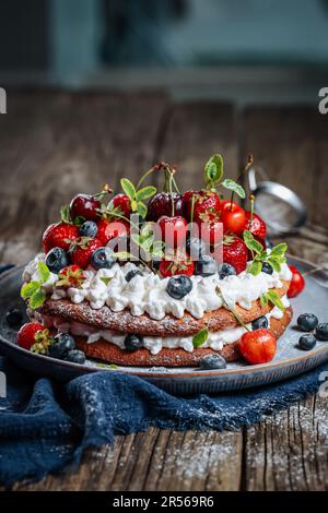 Frischer, saisonaler Kuchen mit Frühlingsreifen Früchten wie Erdbeeren, Heidelbeeren und Kirschen. Auf einem eleganten blauen Teller auf einem rustikalen Holzbrett Stockfoto