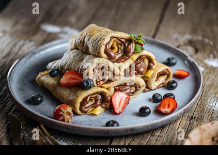 Crepes mit Schokoladenfüllung in Pyramidenform mit Erdbeeren der Saison, Kirschen und Blaubeeren auf einem rustikalen blauen Teller Stockfoto