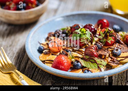 Kleine amerikanische Pfannkuchen mit Obst der Saison wie Erdbeeren, Heidelbeeren und Kirschen. Mit geschmolzener Schokolade getaucht, auf rustikalem Holz Stockfoto