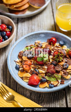 Kleine amerikanische Pfannkuchen mit Obst der Saison wie Erdbeeren, Heidelbeeren und Kirschen. Mit geschmolzener Schokolade getaucht, auf rustikalem Holz Stockfoto