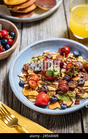 Kleine amerikanische Pfannkuchen mit Obst der Saison wie Erdbeeren, Heidelbeeren und Kirschen. Mit geschmolzener Schokolade getaucht, auf rustikalem Holz Stockfoto