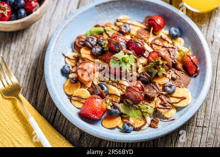 Kleine amerikanische Pfannkuchen mit Obst der Saison wie Erdbeeren, Heidelbeeren und Kirschen. Mit geschmolzener Schokolade getaucht, auf rustikalem Holz Stockfoto