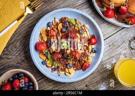 Kleine amerikanische Pfannkuchen mit Obst der Saison wie Erdbeeren, Heidelbeeren und Kirschen. Mit geschmolzener Schokolade getaucht, auf rustikalem Holz Stockfoto