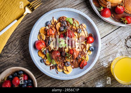 Kleine amerikanische Pfannkuchen mit Obst der Saison wie Erdbeeren, Heidelbeeren und Kirschen. Mit geschmolzener Schokolade getaucht, auf rustikalem Holz Stockfoto