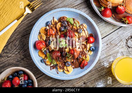 Kleine amerikanische Pfannkuchen mit Obst der Saison wie Erdbeeren, Heidelbeeren und Kirschen. Mit geschmolzener Schokolade getaucht, auf rustikalem Holz Stockfoto