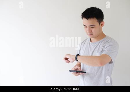 Ein junger asiatischer Mann, der ein graues T-Shirt trägt, hält ein Smartphone und sieht auf eine Smartwatch in der linken Hand. Isolierter weißer Hintergrund. Stockfoto