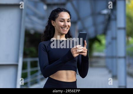 Junge schöne Frau, die telefoniert, eine lächelnde Sportlerin, die sich nach dem Laufen und Sport ausruht, eine lateinamerikanische Frau mit lockigem Haar im Trainingsanzug. Stockfoto