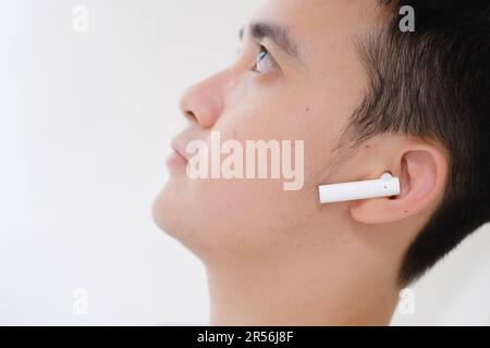 Nahaufnahme des Teilgesichts eines jungen Asiaten, der weiße, echte, kabellose Stereokopfhörer hört. Konzentrieren Sie sich auf den Kopfhörer. Stockfoto