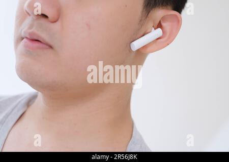 Nahaufnahme des Teilgesichts eines jungen Asiaten, der ein graues T-Shirt trägt und weiße, echte, kabellose Stereo-Ohrhörer hört Stockfoto