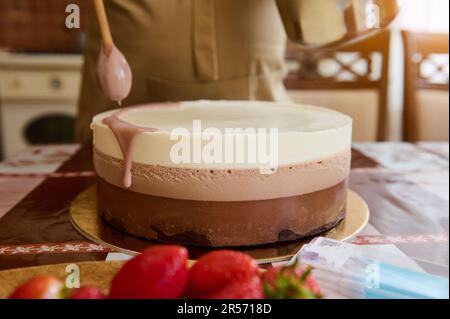 Nahaufnahmen von Konditorin, Konditor gießt rubinrosa Schokoladenglasur auf einen dreifachen Schokoladen-Mousse-Kuchen Stockfoto