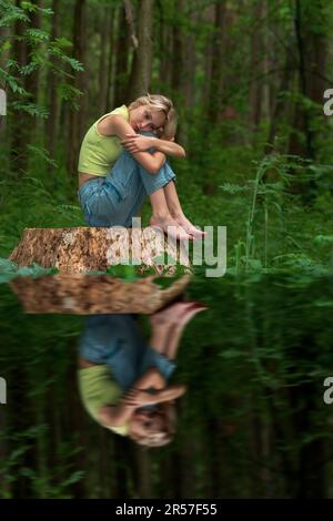 Teenager sitzt auf einem Baumstumpf in einem Naturwaldpark mit einem traurigen und durchdachten Look, der sich im Wasser widerspiegelt Stockfoto