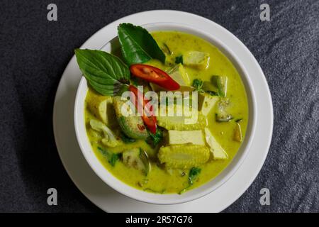 Grüne Curry-Tofu-Suppe. Grüne thailändische Currysuppe in einer weißen Schüssel auf einem weißen Teller mit dunklem Hintergrund. Flach liegend, über Kopf Stockfoto