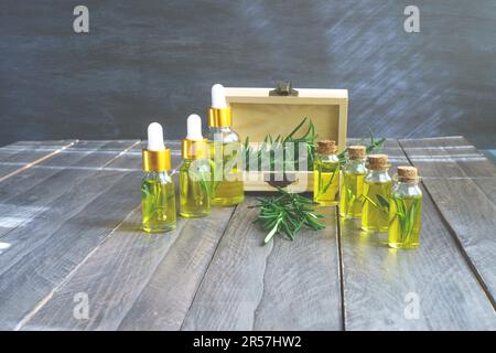 Flaschen in einer Reihe mit Tropfern aus ätherischem Rosmarinöl, einer Holzbox und frischen Rosmarinzweigen, das Sonnenlicht kommt durch das Fenster Stockfoto