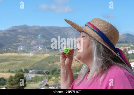 Ältere weißhaarige Frau mit Hut im Profil, die Seifenblasen bläst, mit einer Berglandschaft im Hintergrund Stockfoto