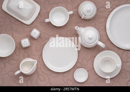 Weißes Porzellan-Geschirr zum Servieren und Essen von Mahlzeiten. Leere saubere Teller, Tasse, Teekanne, Zuckerdose, Milchkanne und Butterschale auf einem Tisch mit beigefarbenem fa Stockfoto