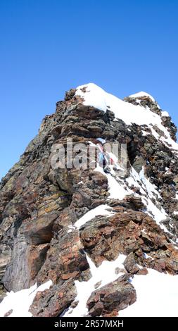 Ein männlicher Backcountry-Skifahrer, der auf einen freiliegenden Felsgipfel klettert, mit seinen Skiern an seinem Rucksack in den Silvretta-Bergen an der Grenze zu Stockfoto