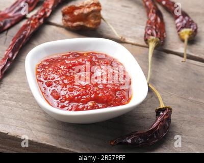 Sehr würzige Chilisauce in einer weißen Schüssel auf einem rustikalen Holzküchentisch, getrocknete Chilischoten als Dekoration Stockfoto