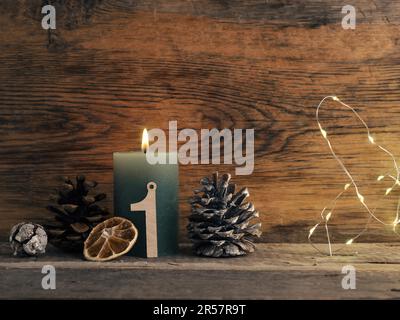 Kerze vom ersten Advent brennt, Tannenzapfen und Weihnachtsdekoration auf Holz mit Weihnachtslichtern Stockfoto