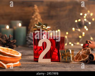 Kerze vom zweiten Advent brennt, Holznummer zwei mit Geschenkschachteln, Tannenzapfen und Weihnachtsdekoration auf Holz mit Weihnachtslichtern Stockfoto