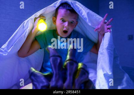 Ein verängstigter Junge mit Taschenlampe versteckt sich unter der Decke vor der Hand eines gruseligen Monsters Stockfoto