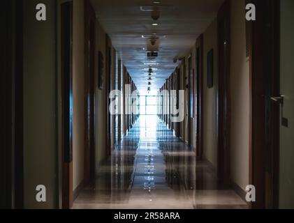 Dunkler langer Korridor im Hotel mit vielen Türen und Licht am Ende durch das große Fenster. Stockfoto
