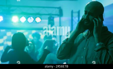 Männlicher Erwachsener, der auf einer Party telefoniert, versucht, mit jemandem über ein Handy zu reden, Ferngespräch in der Diskothek. Junge Person, die auf einer Tanzparty im Club ist, geht ans Smartphone. Stativaufnahme. Stockfoto