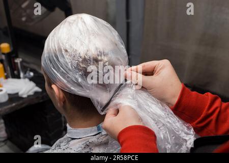Friseurhände Nahaufnahme Fixieren einer Schutzfolie auf dem Kopf eines Klienten, das Färben von Haaren in einem Friseursalon. Stockfoto