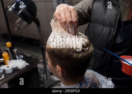 Die Hand der Freundin eines professionellen Friseurs berührt und färbt das Haar des Freundes des Kunden in einem Friseur oder Friseursalon weiß. Stockfoto