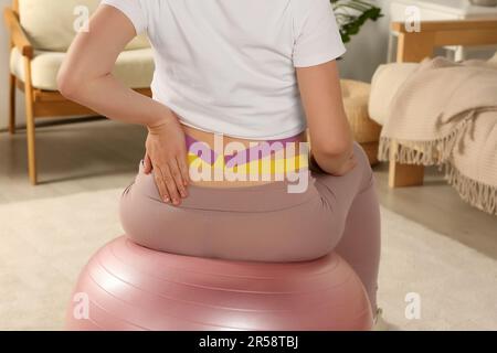 Nahaufnahme einer schwangeren Frau mit Kinesiobändern, die zu Hause Übungen am Sportball macht, Rückansicht Stockfoto