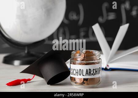 Stipendienkonzept. Glasbecher mit Münzen und Graduierungskappe auf weißem Holztisch Stockfoto