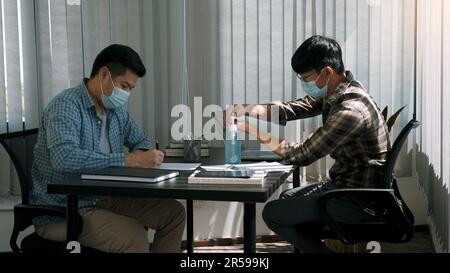 Asiatische Kollegen arbeiten im Büro, während sie eine Maske tragen und während des Virenausbruchs Handdesinfektionsmittel verwenden. Stockfoto