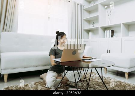 Junge asiatische Frau sitzt im Wohnzimmer mit einem Taschenrechner, um Familieneinkommen und Ausgaben zu berechnen und schreibt es in seinem Notebook oder Laptop auf h Stockfoto