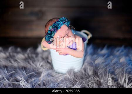 Nahaufnahmen eines neugeborenen Mädchens in einem weißen Metallwürfel, das eine riesige Brille und ein blaues Blumenband trägt. Stockfoto