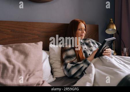 Seitenansicht einer trauernden jungen Frau, die auf dem Bett liegt, ein Foto im Rahmen hält und mit geschlossenen Augen weint, das Bild zu Hause mit Liebe berührt. Stockfoto