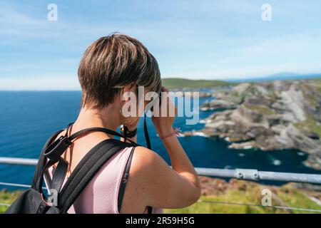 Ein weißer Tourist fängt Irlands atemberaubende Schönheit ein, während sie üppige grüne Klippen in bezauberndem Licht unter einem ruhigen azurblauen Himmel fotografiert Stockfoto