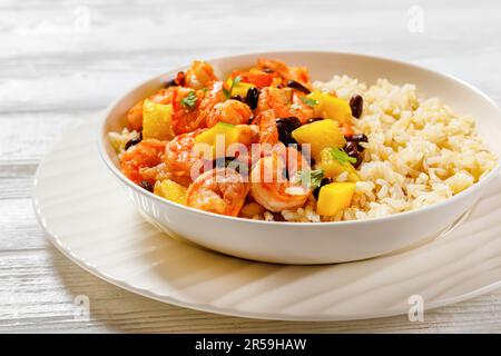 Karibische gegrillte Garnelen mit schwarzen Bohnen, Mango-Ananassalsa serviert mit braunem Reis in einer weißen Schüssel auf einem strukturierten Holztisch, Landschaftsblick Stockfoto