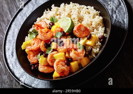 Karibische gegrillte Garnelen mit schwarzen Bohnen, Mango-Ananassalsa serviert mit braunem Reis in schwarzer Schüssel auf dunklem Holztisch, Nahaufnahme Stockfoto