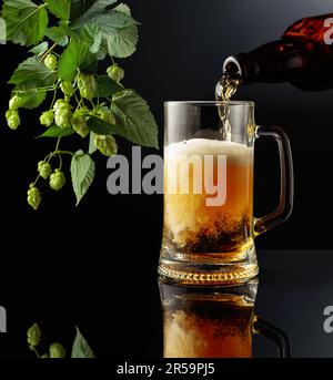 Bier aus einer Flasche in eine Tasse gießen. Bierbecher und Hopfenzweig auf schwarzem reflektierendem Hintergrund. Speicherplatz kopieren. Stockfoto