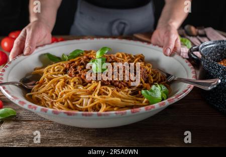 Frau, die eine große Nudelschüssel mit Spaghetti Bolognese auf rustikalem Holztisch hält oder serviert Stockfoto