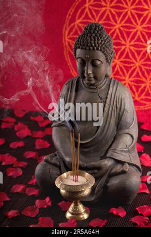 Buddha mit Räucherstäbchen unter Blütenblättern und Blume des Lebens im Hintergrund Stockfoto