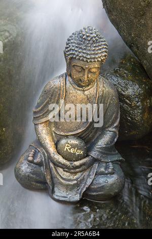 buddha-Statue in einem Wasserfall, in den Händen eine Steinschrift ENTSPANNT Stockfoto