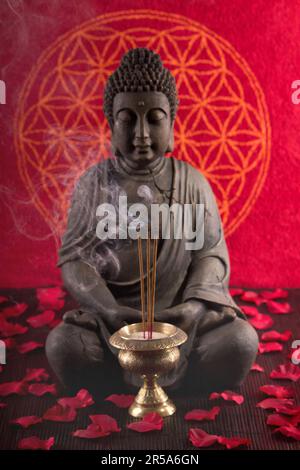 Buddha mit Räucherstäbchen unter Blütenblättern und Blume des Lebens im Hintergrund Stockfoto