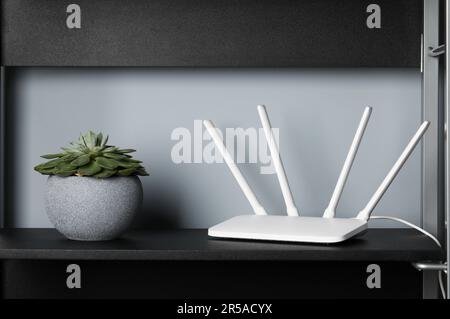 Neuer weißer WLAN-Router in der Nähe einer Topfpflanze auf schwarzem Regal Stockfoto