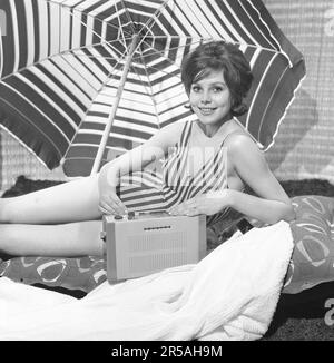 1960er Sommer-Lifestyle. Eine junge Frau in einem Fotografen-Studio, die einen Badeanzug mit einem tragbaren Radio trägt. Der Sonnenschirm im Hintergrund ist dort platziert, um das Sommergefühl zu verstärken. Schweden 1960 Photo Kristoffersson Ref CL114-10 Stockfoto