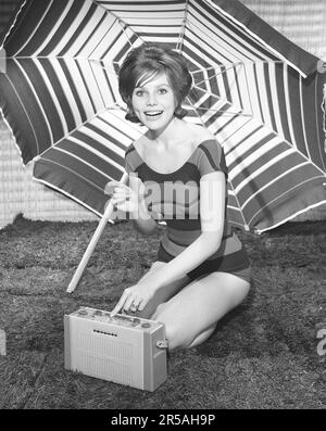 1960er Sommer-Lifestyle. Eine junge Frau in einem Fotografen-Studio, die einen Badeanzug mit einem tragbaren Radio trägt. Der Sonnenschirm im Hintergrund ist dort platziert, um das Sommergefühl zu verstärken. Schweden 1960 Photo Kristoffersson Ref CL113-4 Stockfoto